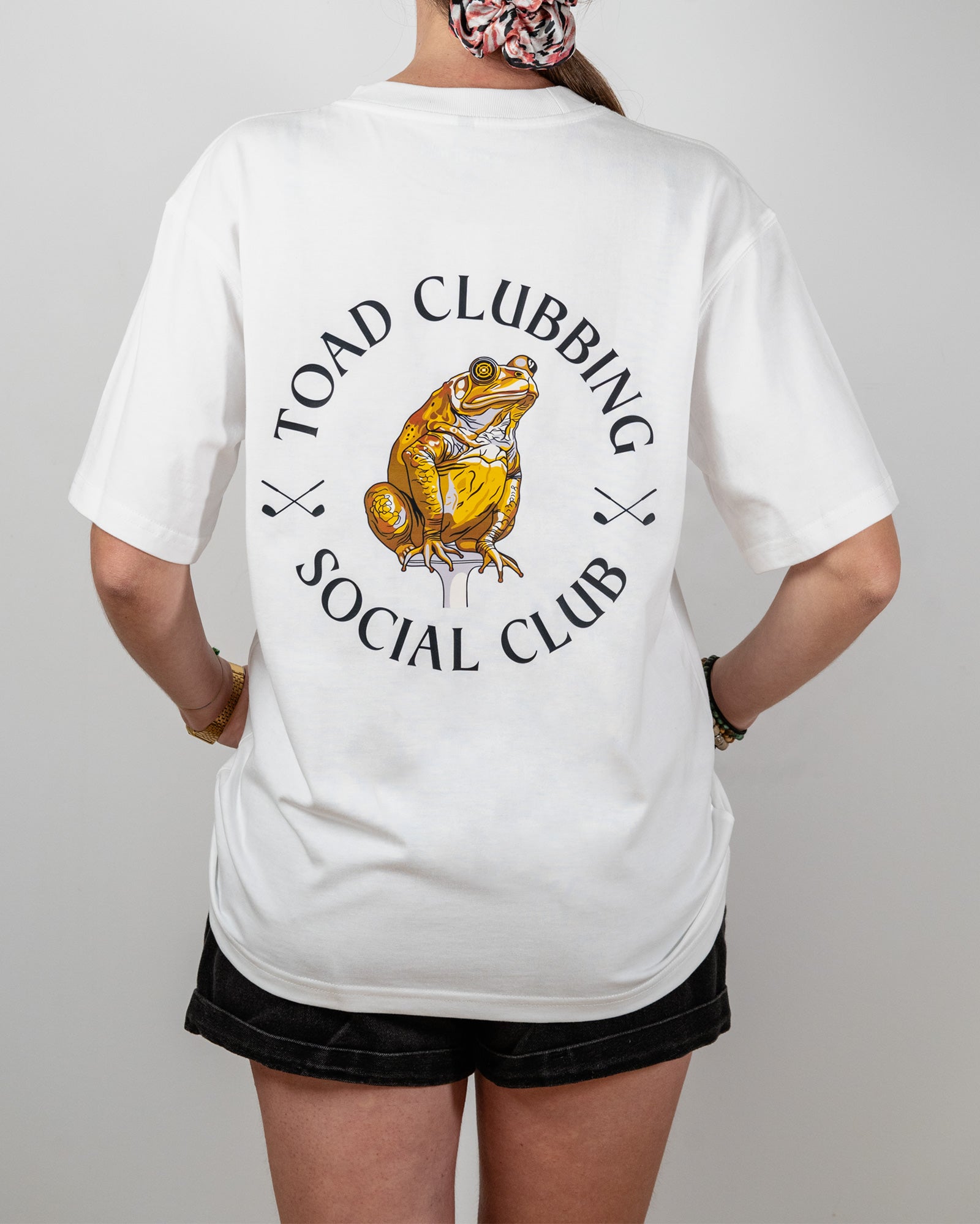 Person wearing a white t-shirt with a graphic of a toad and text 'Toad Clubbing Social Club' on the back.