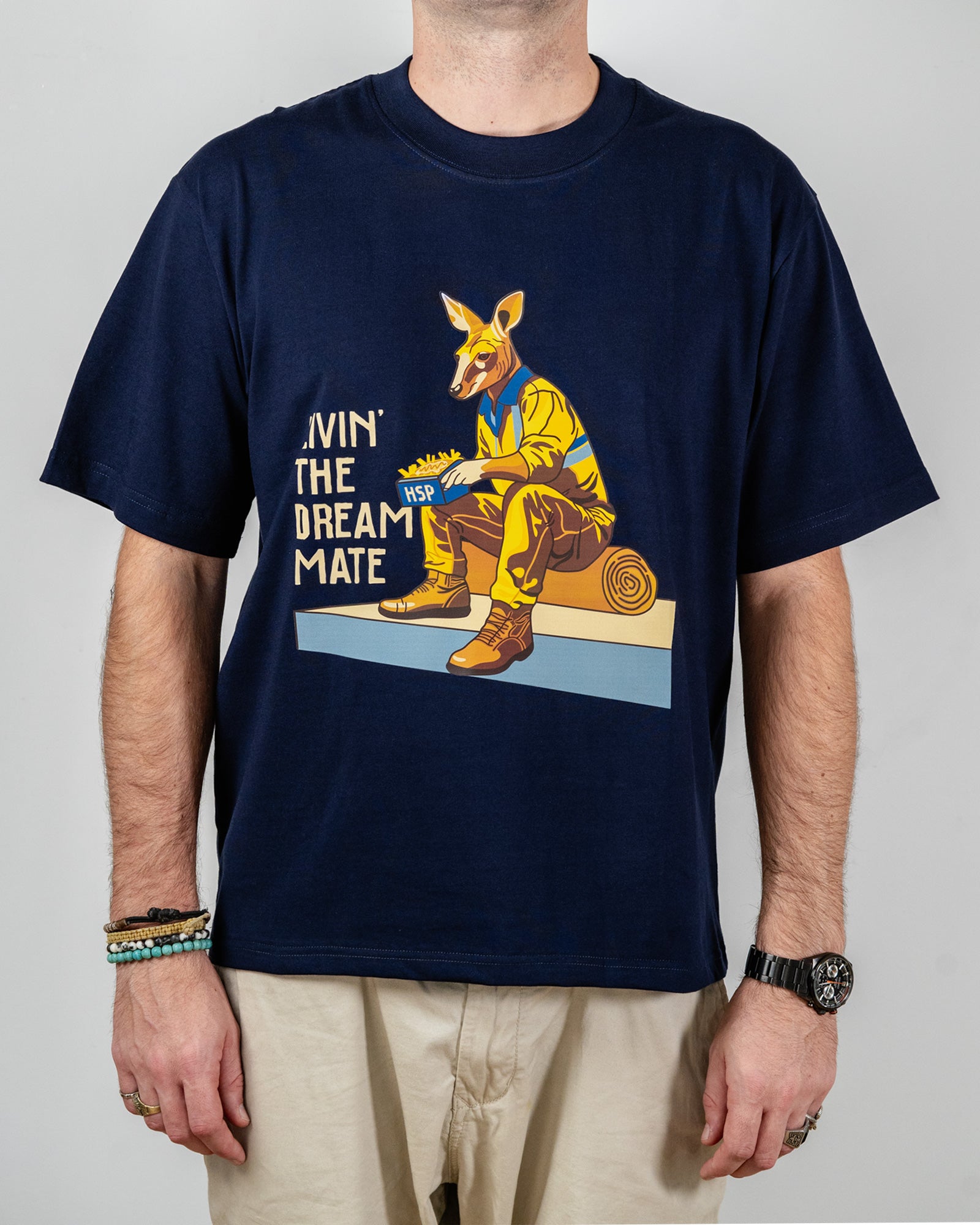 Navy blue t-shirt with a "Livin' The Dream Mate" graphic —featuring a kangaroo wearing a hi-vis set of overalls and work boots, eating an HSP kebab pack whilst sitting on a wooden log that is on top of a concrete slab.