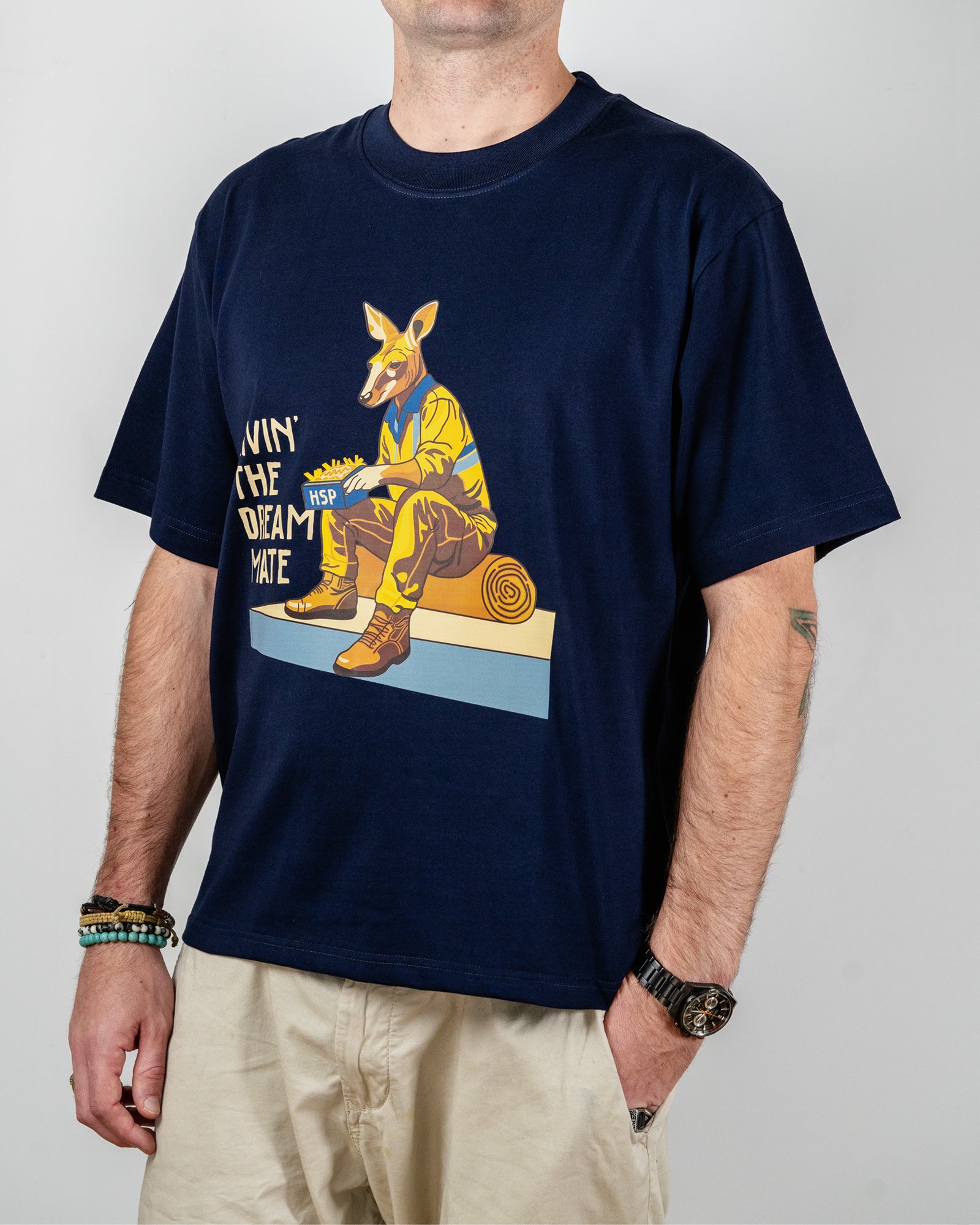 Navy blue t-shirt with a "Livin' The Dream Mate" graphic —featuring a kangaroo wearing a hi-vis set of overalls and work boots, eating an HSP kebab pack whilst sitting on a wooden log that is on top of a concrete slab.