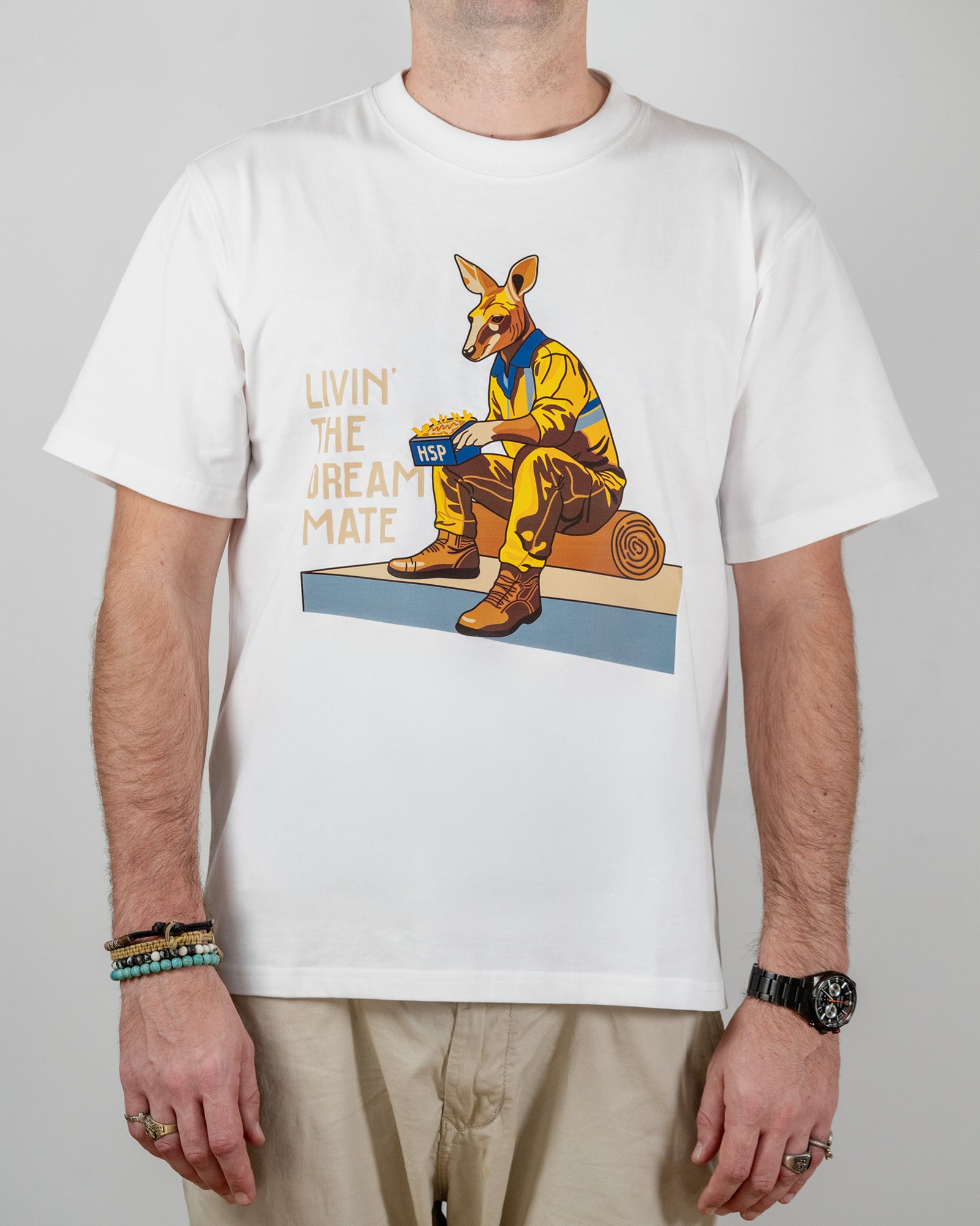 White t-shirt with a "Livin' The Dream Mate" graphic —featuring a kangaroo wearing a hi-vis set of overalls and work boots, eating an HSP kebab pack whilst sitting on a wooden log that is on top of a concrete slab.