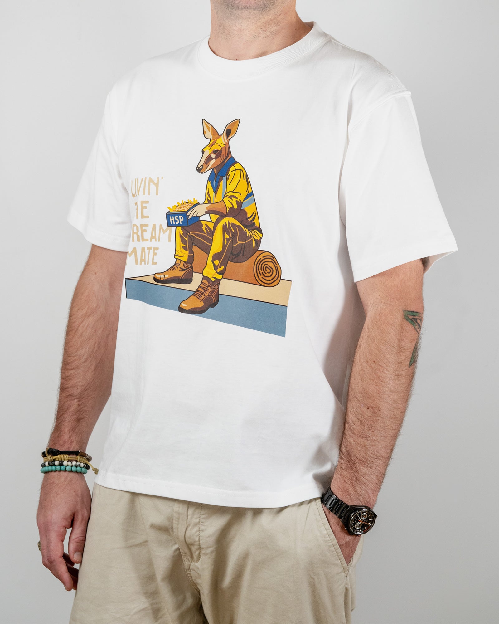 White t-shirt with a "Livin' The Dream Mate" graphic —featuring a kangaroo wearing a hi-vis set of overalls and work boots, eating an HSP kebab pack whilst sitting on a wooden log that is on top of a concrete slab.
